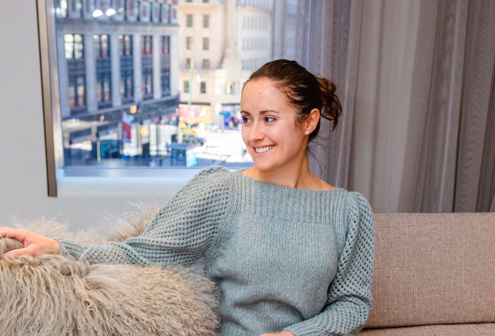 Personal Stylist Dina Scherer sits comfortably on a beige couch, dressed in a light blue knitted sweater. Her smile radiates warmth as she glances slightly to her right. Behind her, the cityscape is visible through a large window, creating an inviting blend of urban charm and cozy elegance.