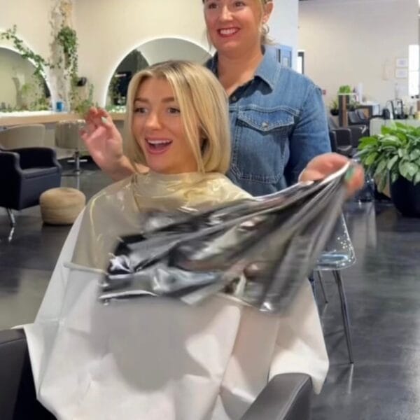 A smiling blonde woman removes a metallic cape in a salon chair, happy with her new haircut after receiving the Color Analysis + Facial Features Assessment from an Image and Wardrobe Stylist Sarasota in a denim shirt.