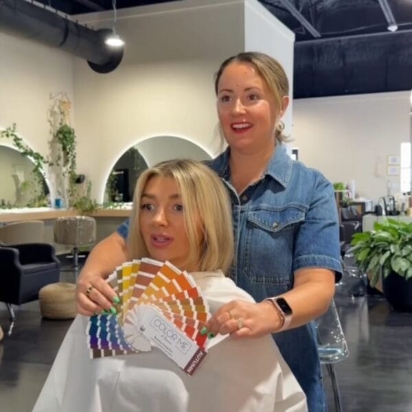 A hairdresser stands behind a client in a modern salon, holding color sample cards for the Color Analysis + Facial Features Assessment. The client in a white cape looks ahead while the smiling stylist conducts the session amid stylish décor and plants.