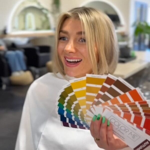 A smiling blonde woman holds colorful hair swatches in a salon, suggesting she’s choosing a new look—perhaps after a Color Analysis + Facial Features Assessment with an Image and Wardrobe Stylist Sarasota. Salon chairs and mirrors fill the background.