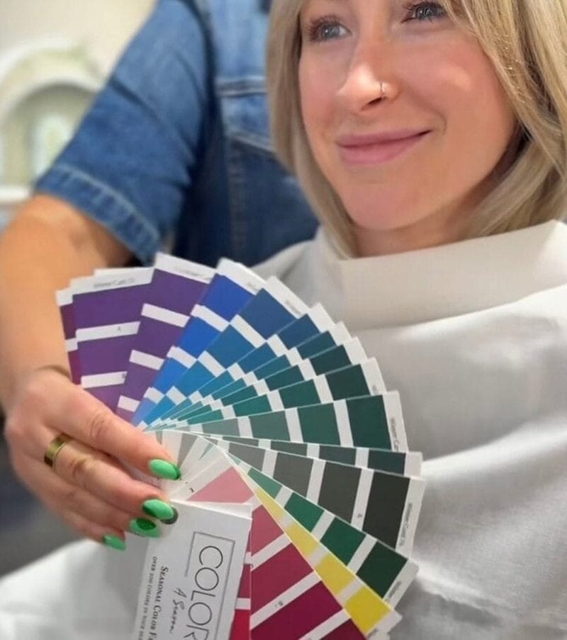 A smiling blonde woman in a salon cape holds colorful swatches during a Color Analysis + Facial Features Assessment, while an Image and Wardrobe Stylist Sarasota shows the swatches with green-painted nails.