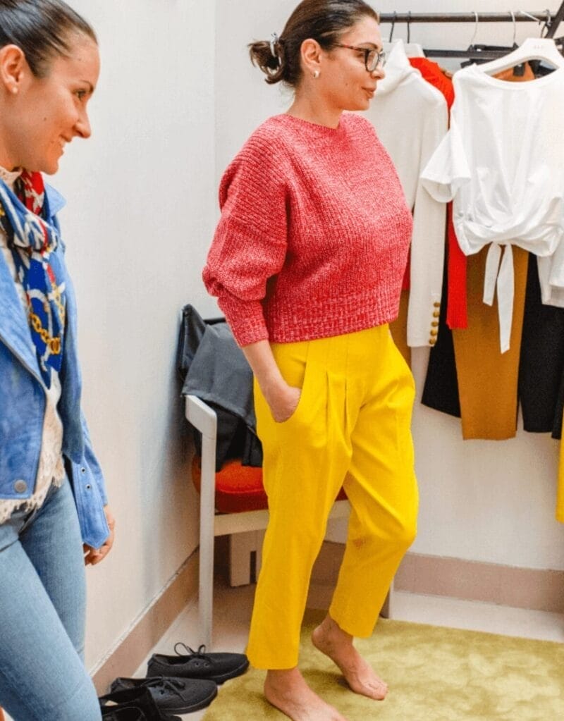 Two women are in a fitting room. One, barefoot, tries on a red sweater and yellow pants as her friend—an Image and Wardrobe Stylist Sarasota—offers advice. Various clothes hang on a rack in the background.