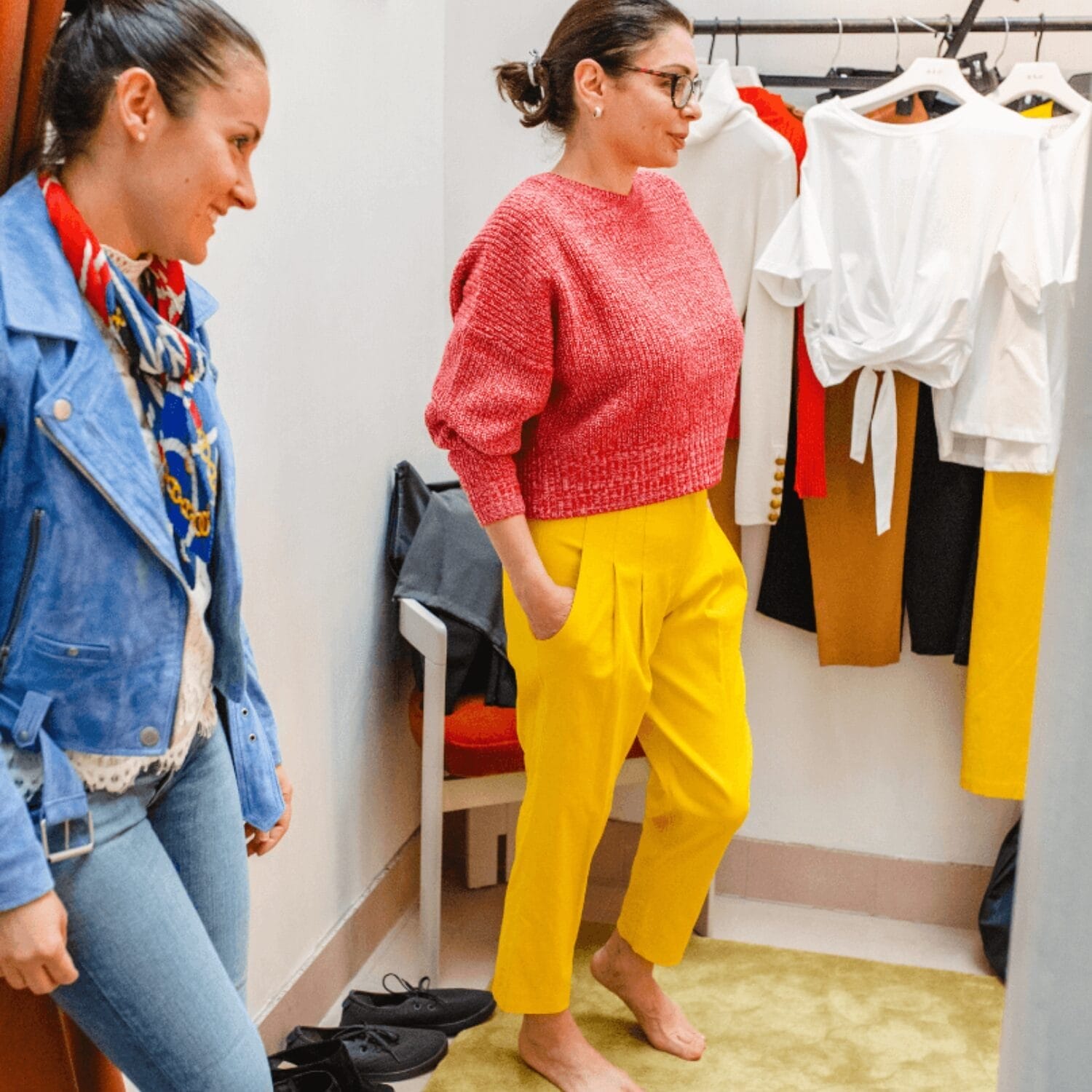 Two women are in a fitting room. One, barefoot, tries on a red sweater and yellow pants as her friend—an Image and Wardrobe Stylist Sarasota—offers advice. Various clothes hang on a rack in the background.