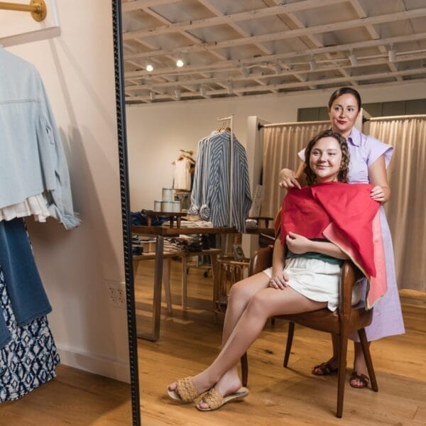 A woman drapes a red cloth over another woman seated in front of a mirror in a stylish clothing store, suggesting a personal shopping or virtual color analysis session. Clothing racks and a wooden floor are visible in the background.