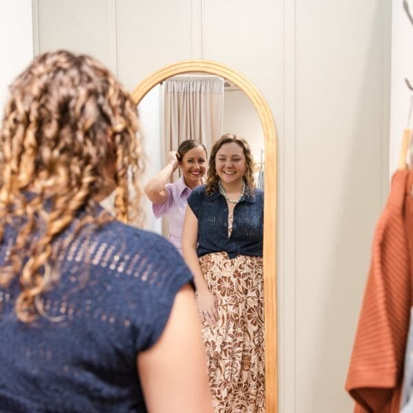 Two women smiling in front of a mirror; one stands behind the other, helping fix her hair. Their happy reflection captures a moment of joy. Clothing and a light-colored wall are visible—perfect for an Image and Wardrobe Stylist Sarasota session.